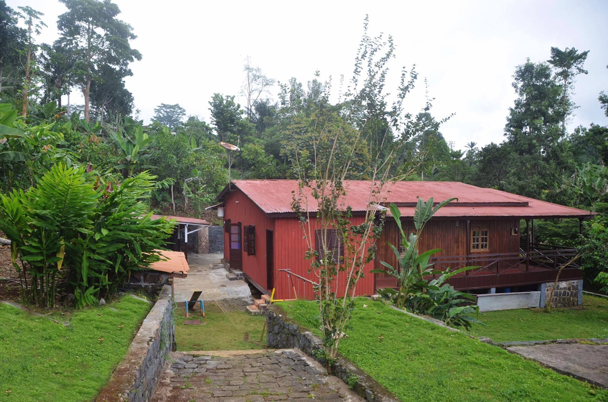 A red wooden house with a red roof, surrounded by lush greenery and a stone pathway leading to the entrance.