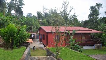 A red wooden house with a red roof, surrounded by lush greenery and a stone pathway leading to the entrance.