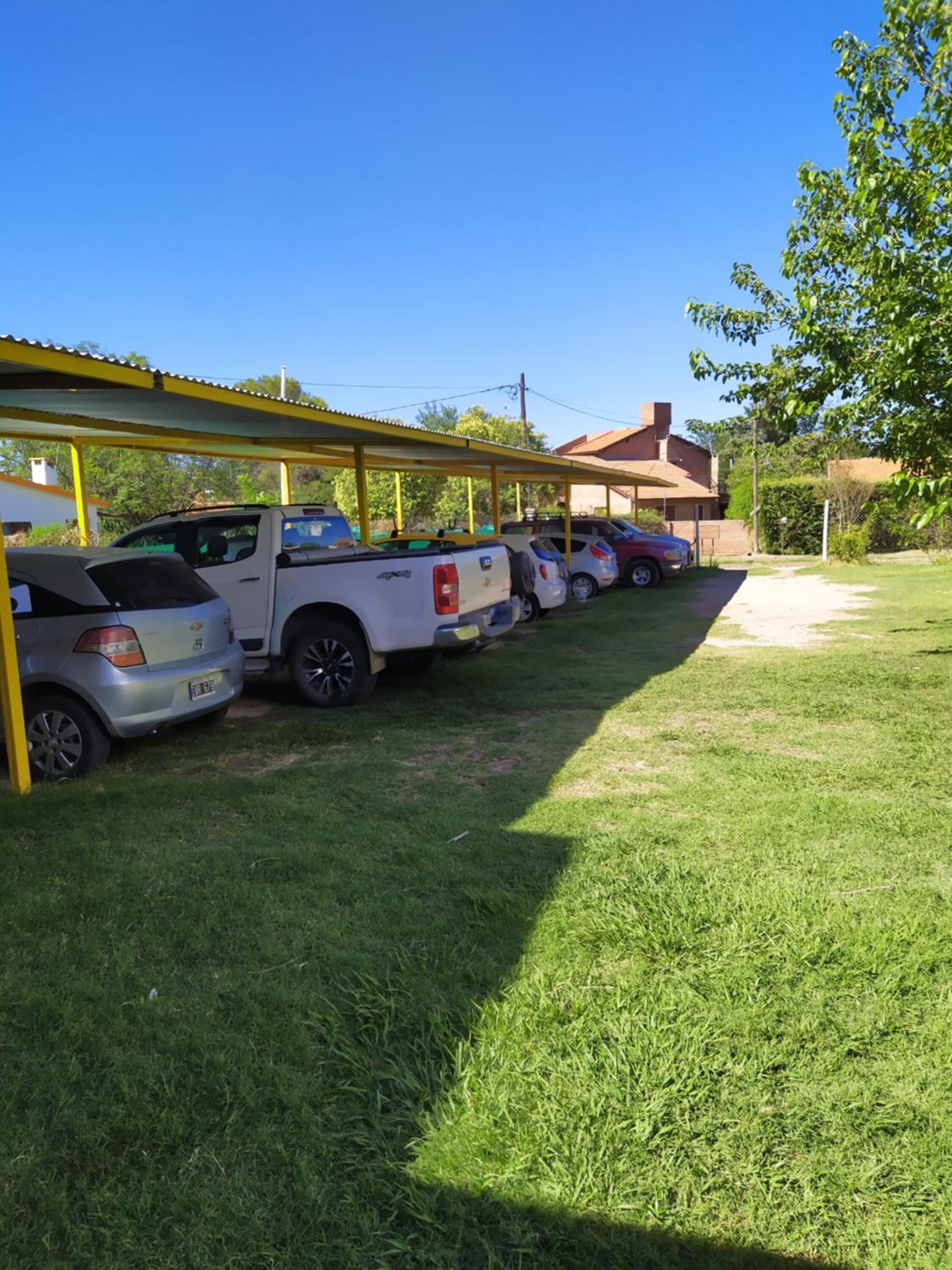 Reihe geparkter Fahrzeuge unter einem überdachten Parkbereich mit einer Rasenfläche im Vordergrund und Gebäuden im Hintergrund.