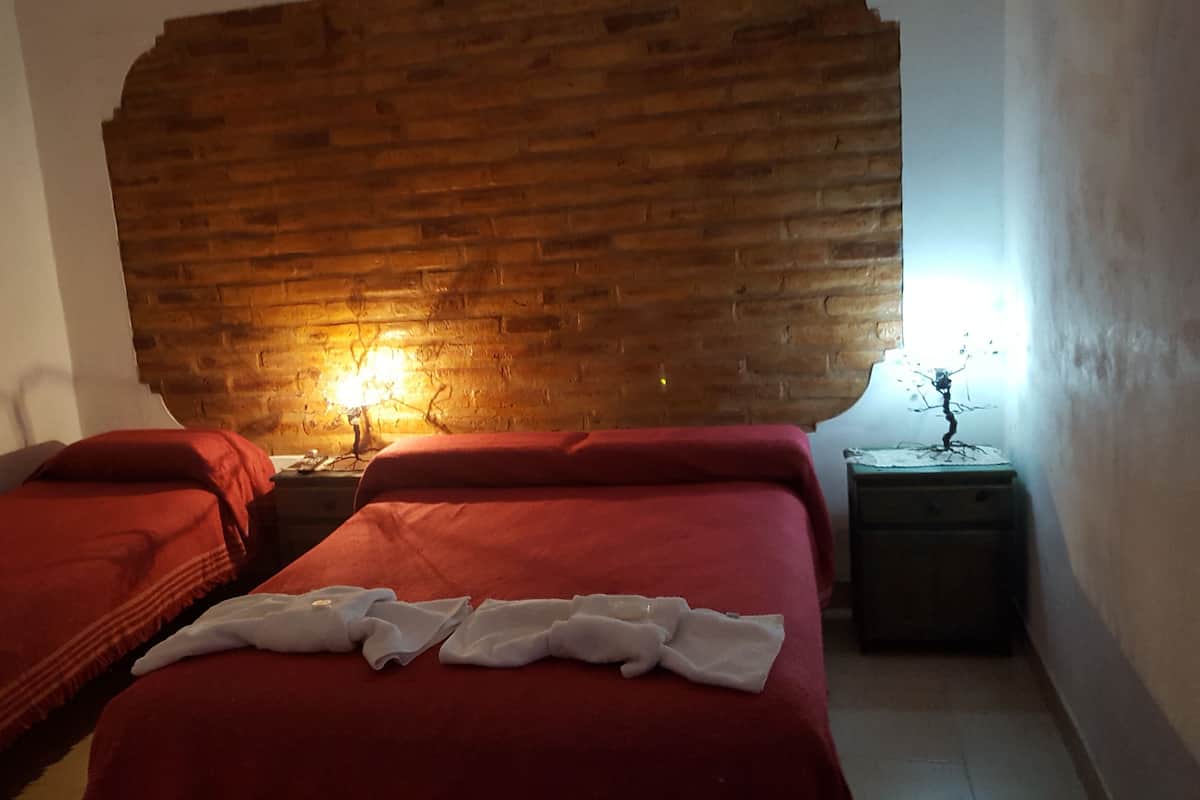 Ein Zimmer mit zwei Betten, jeweils mit roter Bettwäsche und einer weißen Decke. Im Vordergrund befindet sich ein Bett. Dahinter eine Ziegelwand und ein Nachttisch mit einer Lampe zwischen den Betten.