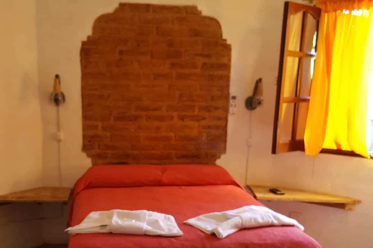 Ein Bett mit roter Bettdecke und weißen Kissen, einem Ziegel-Kopfstück, einer hängenden Leuchte, einem Fenster mit gelben Vorhängen und einem gefliesten Boden.