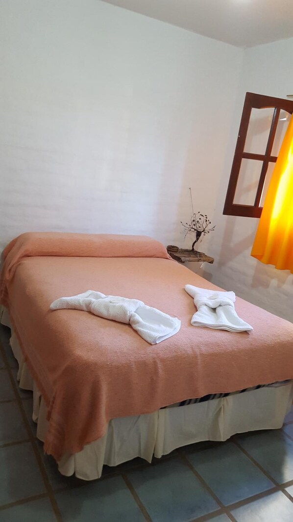 Un lit bien fait, avec une couvre-lit rose et deux serviettes blanches, une petite table de chevet agrémentée d’un objet décoratif, et une fenêtre avec un rideau.