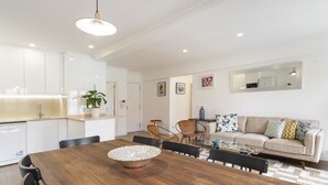 A modern kitchen with white cabinets, a wooden dining table with chairs, a sofa with cushions, and a mirror on the wall.
