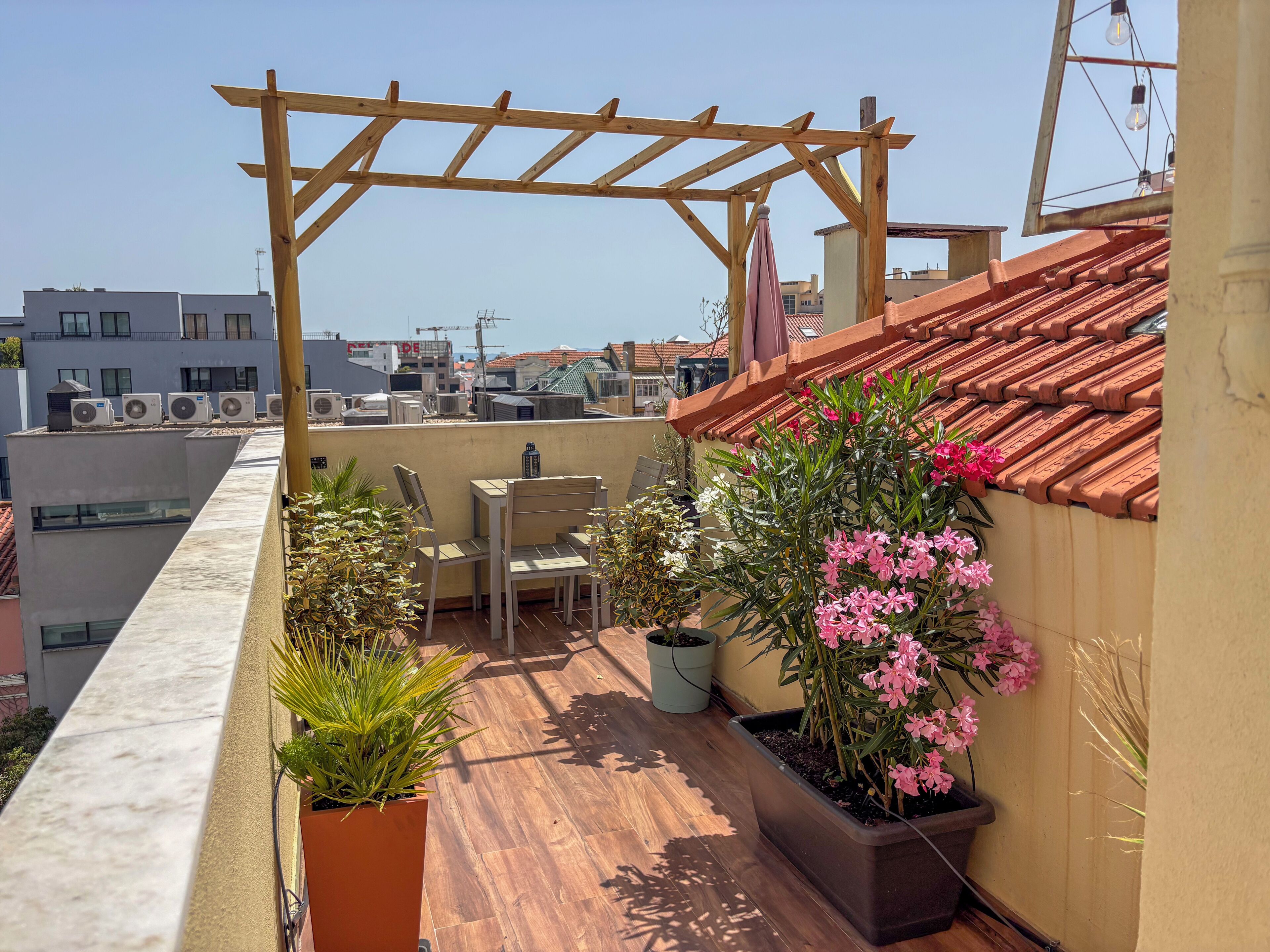 A rooftop terrace with a pergola, seating, potted plants, and a view of surrounding buildings.