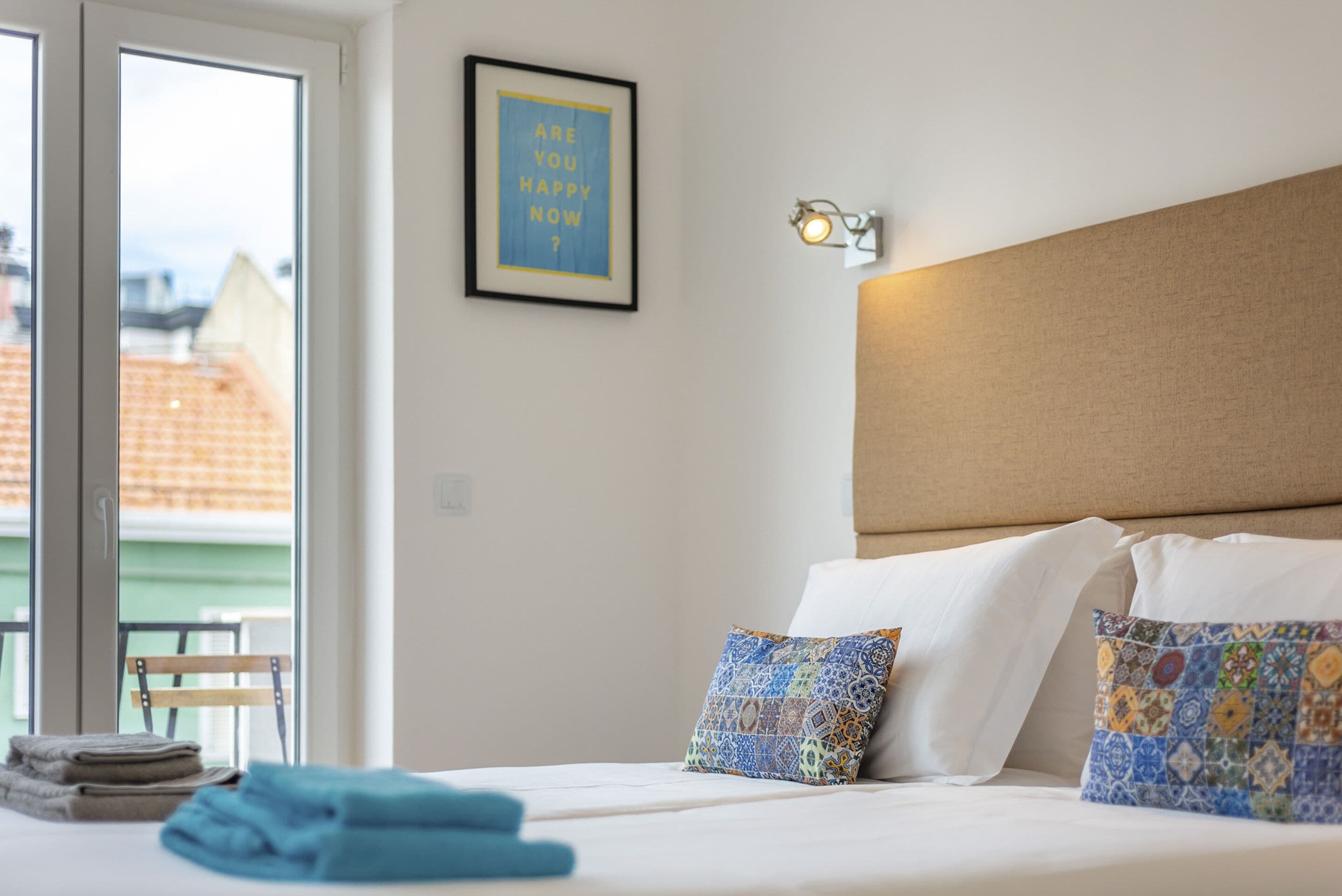 A hotel room with a bed, pillows, a framed quote on the wall, and a window with a view of buildings.
