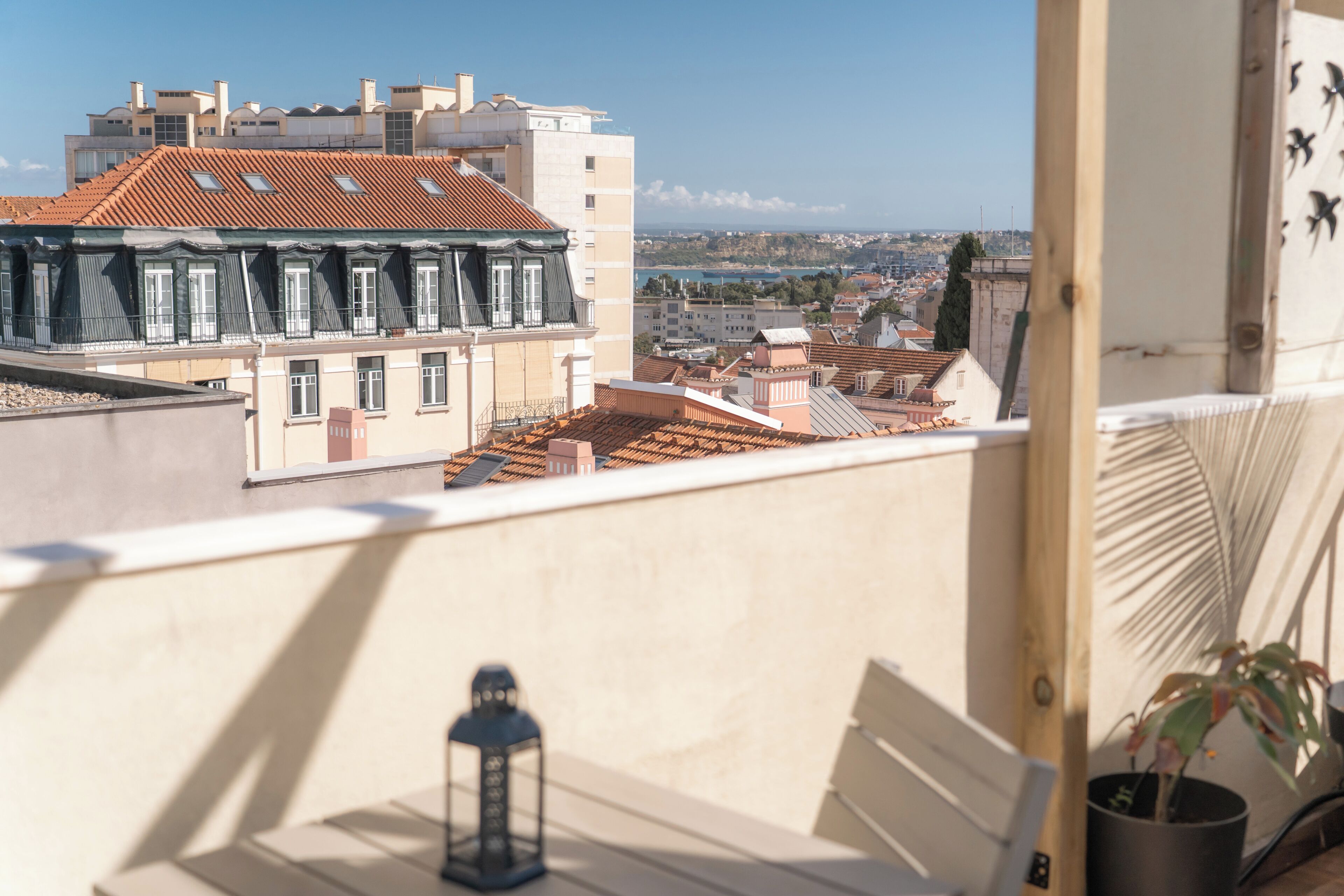 Un balcon offrant une vue sur le paysage urbain, avec des bâtiments aux toits recouverts de tuiles rouges et une étendue d’eau visible au loin.