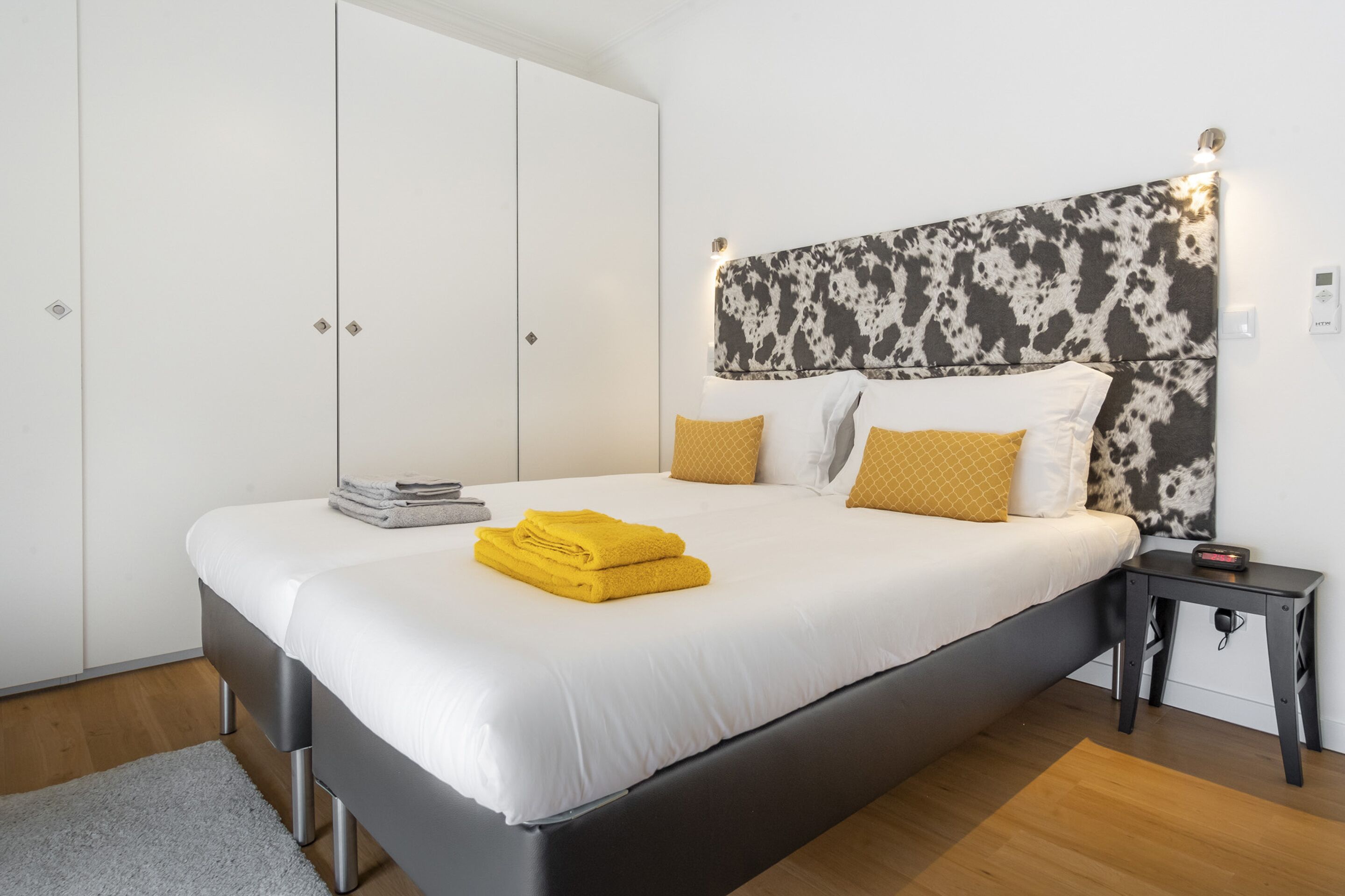 Een net opgemaakt bed met wit en geel beddengoed, een hoofdbord met een zwart-wit patroon en een klein nachtkastje met een lamp.