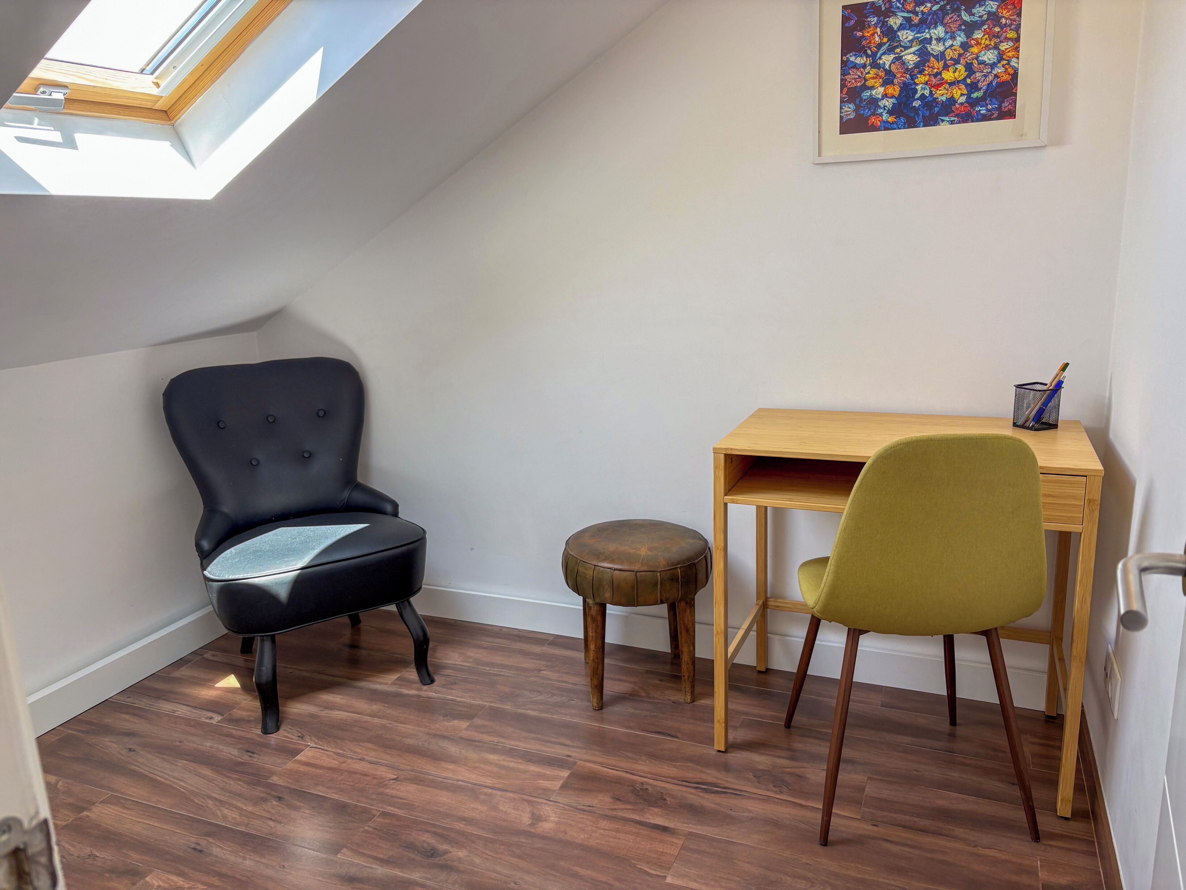 A room with a skylight, a black chair, a wooden desk, a green chair, and a small stool.
