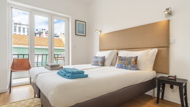 A hotel room with a large bed, two bedside tables, a chair, and a view of buildings through a glass door.