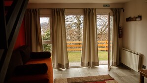 Een kamer met een bank, een houten bankje buiten en uitzicht op bomen en een hek.