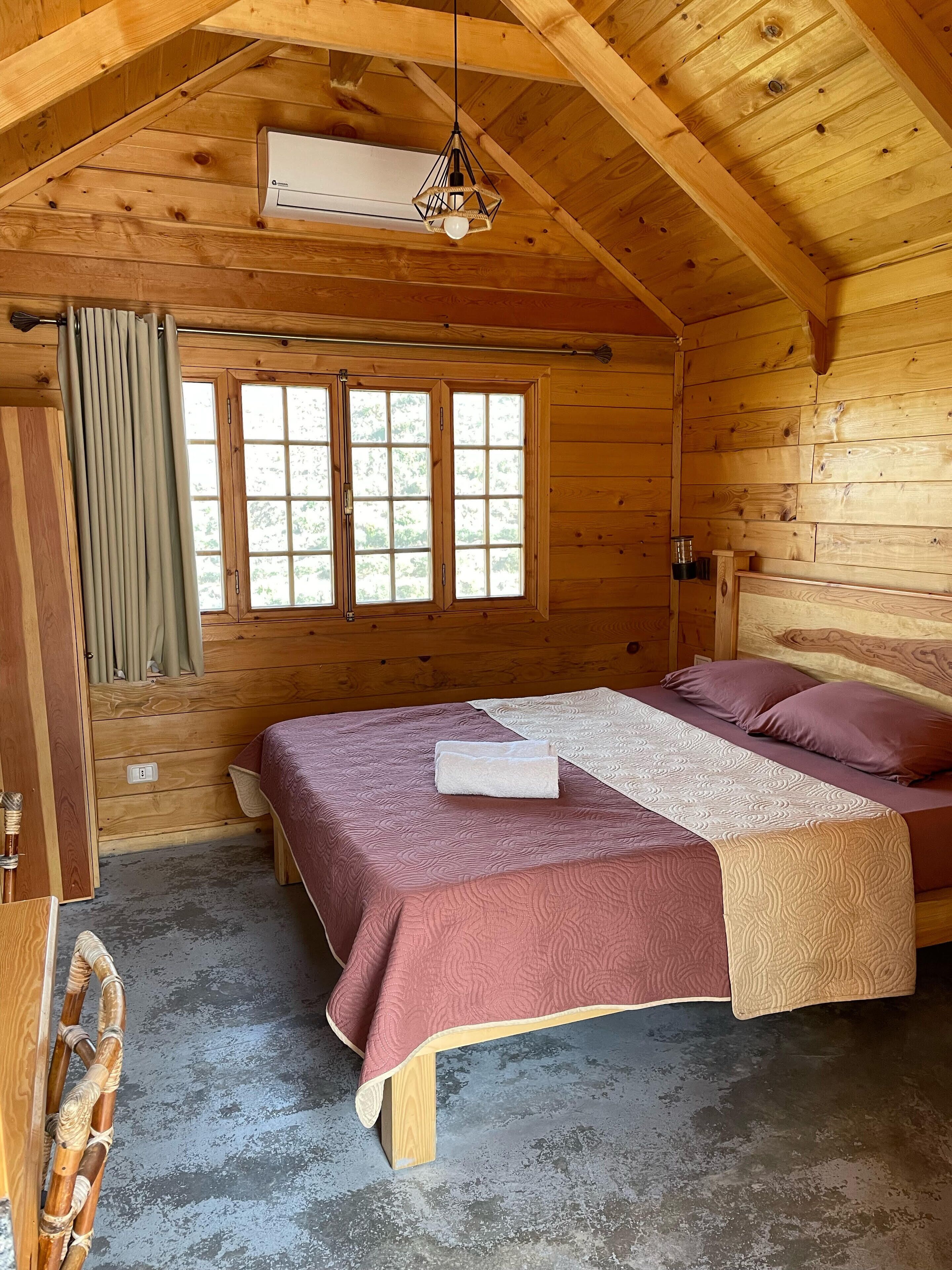 Un dormitorio de cabaña de madera con cama, ventana y lámpara colgante.