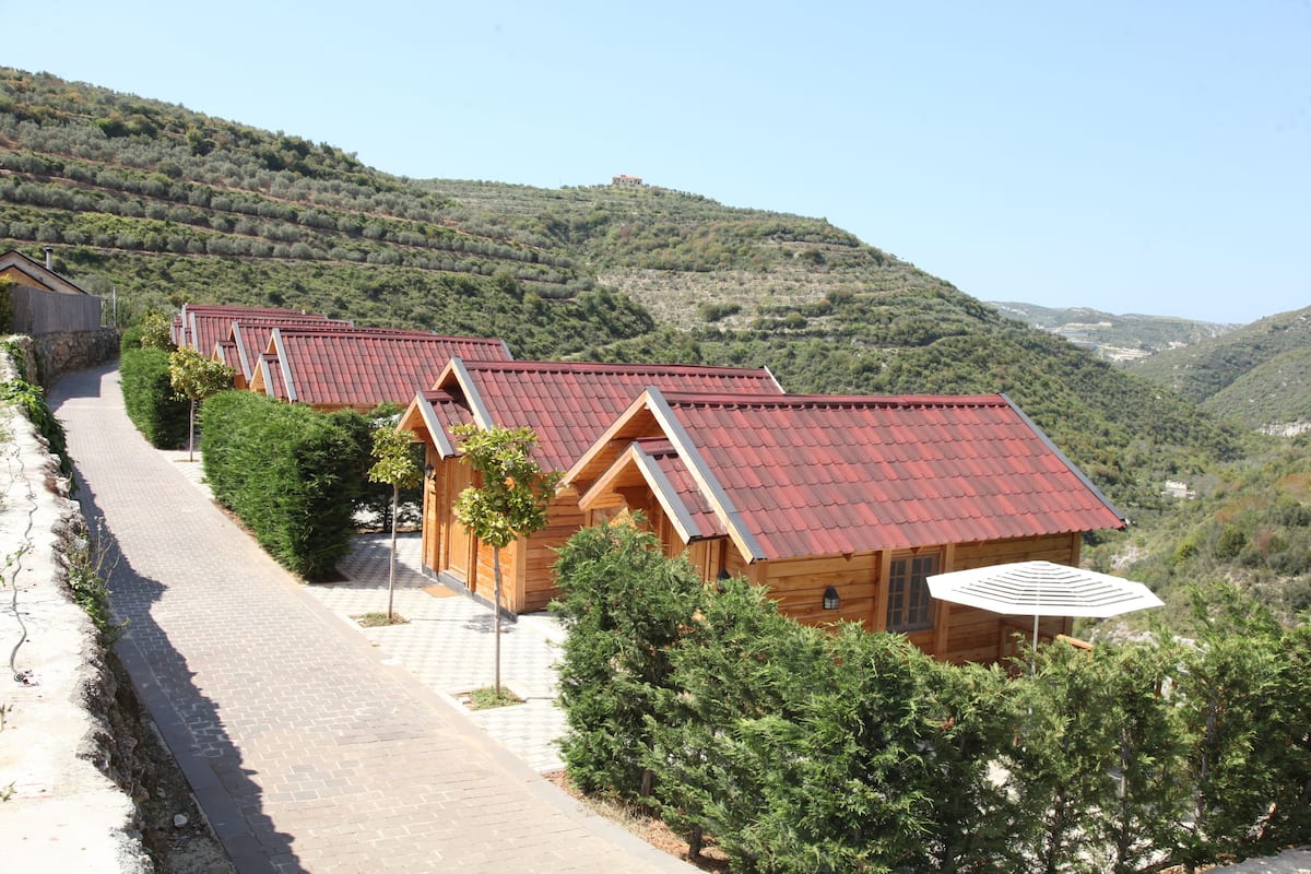 A row of wooden cabins with red roofs, set against a hilly backdrop.
