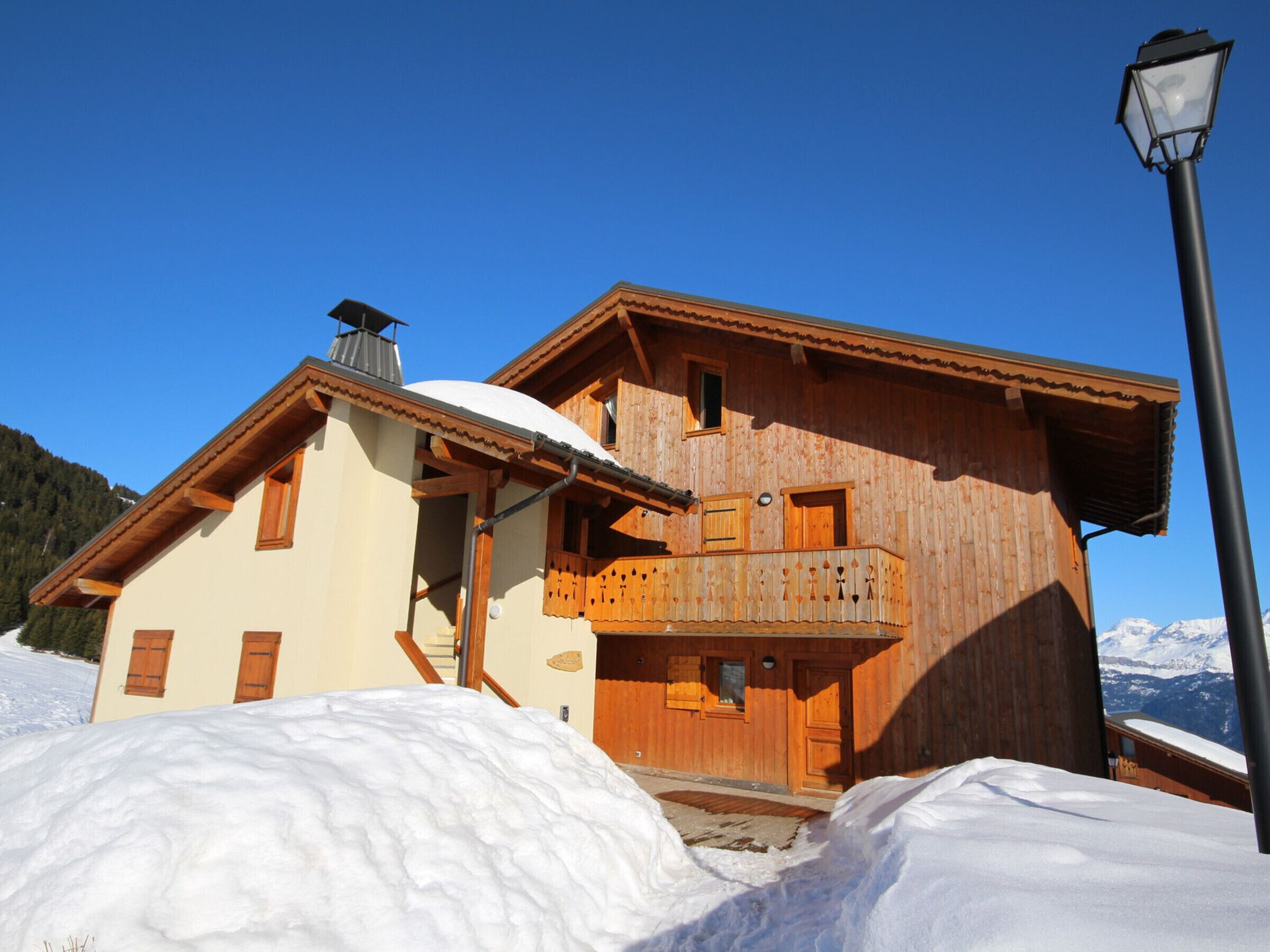 A wooden chalet with a balcony, surrounded by snow, with a clear sky and mountains in the background.
