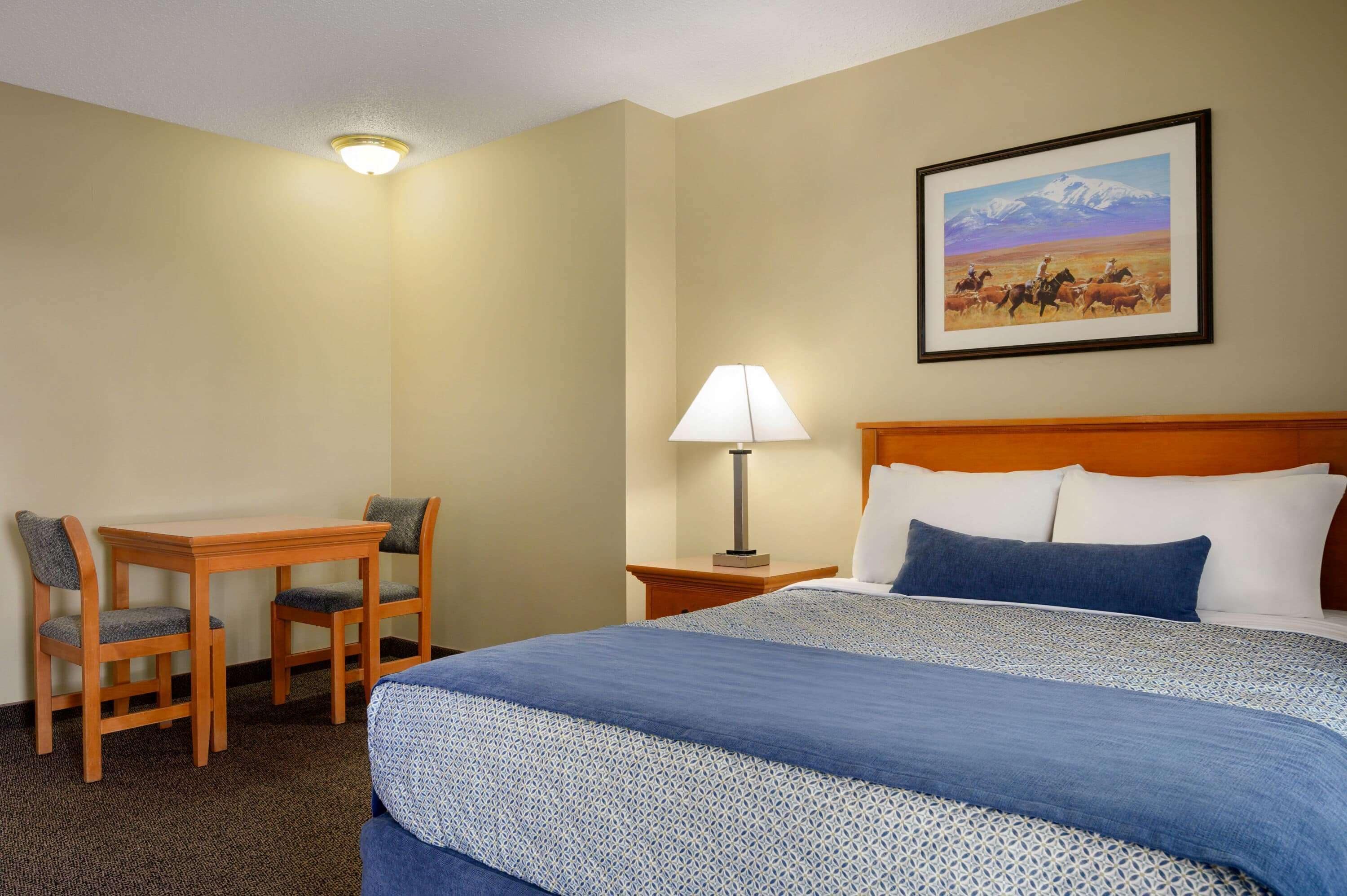 Habitación de hotel con una cama, un escritorio con dos sillas, una lámpara y un cuadro en la pared.