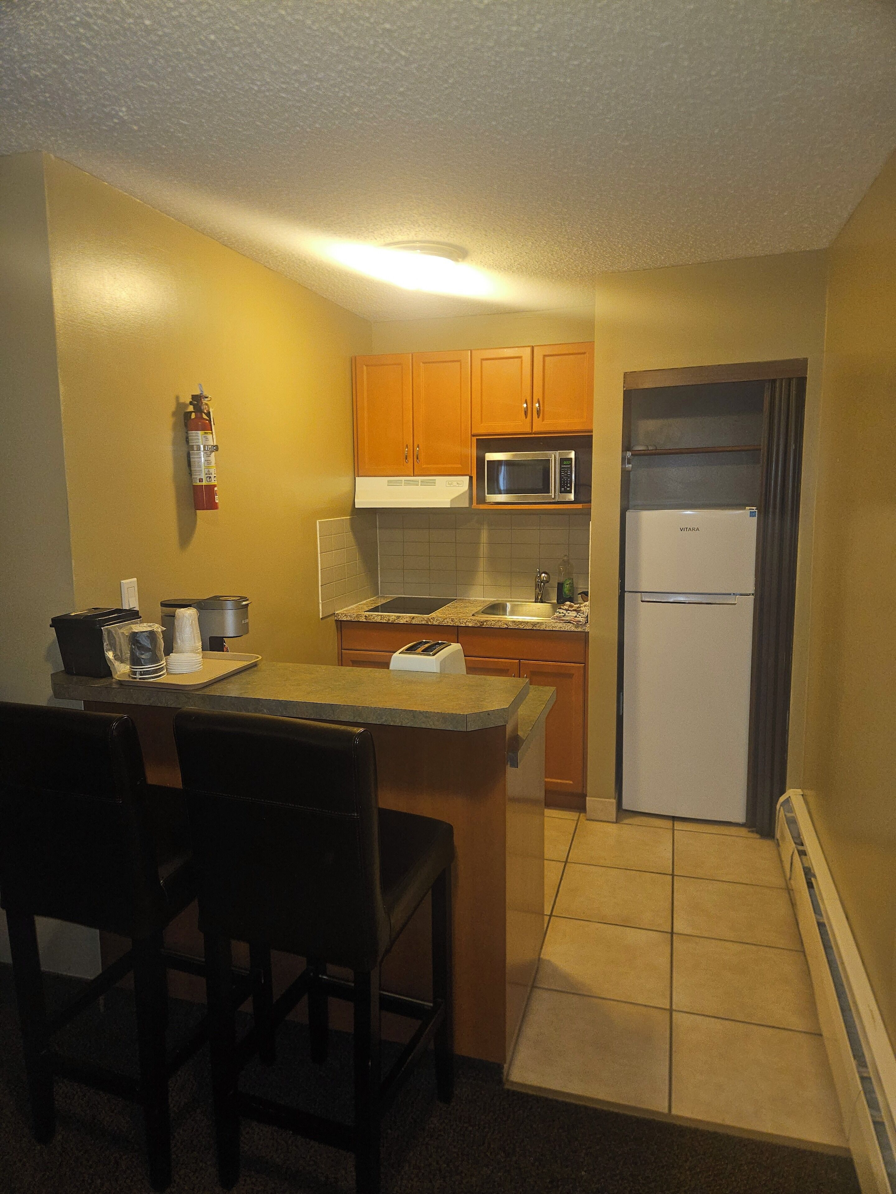 A compact kitchen with wooden cabinets, a microwave, a Vitara refrigerator, a sink, and a small dining area with chairs.