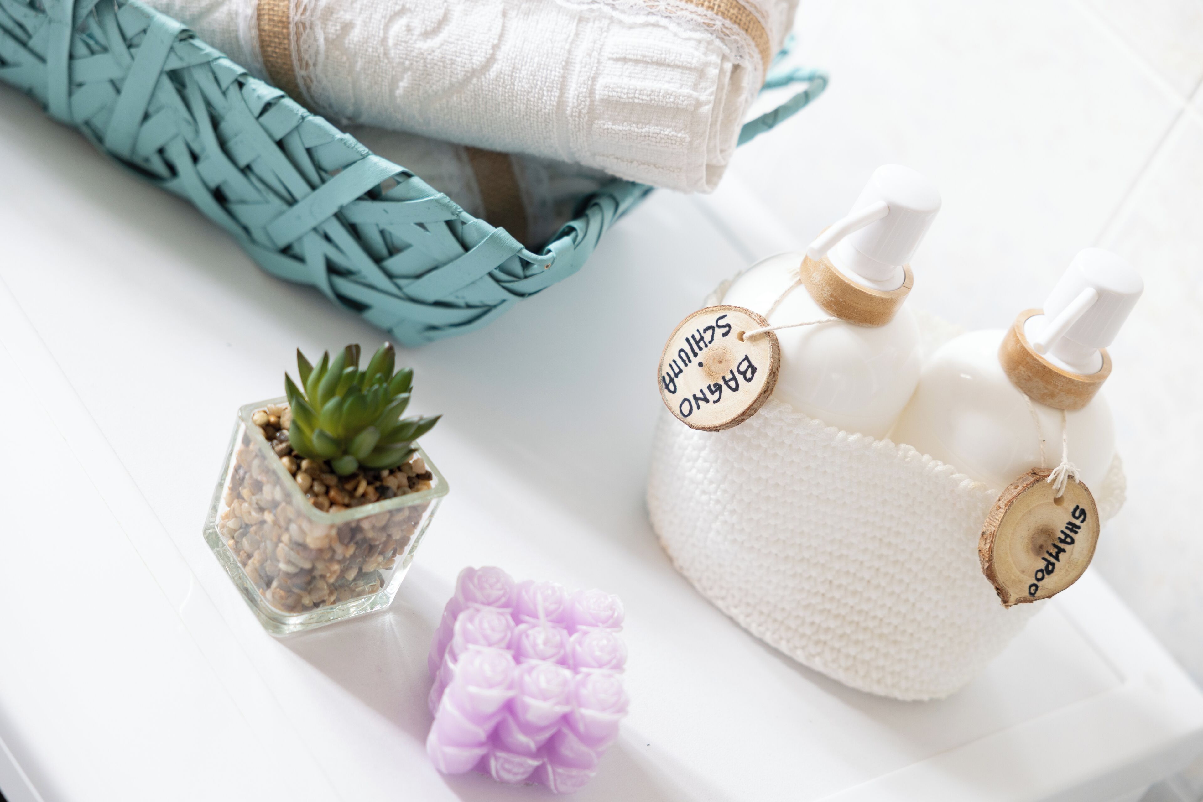 A white surface with a woven blue towel, a small potted succulent, a purple rose-shaped object, and two white bottles with wooden labels.