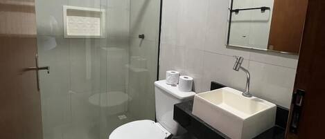 Un baño moderno con ducha de vidrio, inodoro blanco, lavamanos con espejo y una toalla en el suelo.