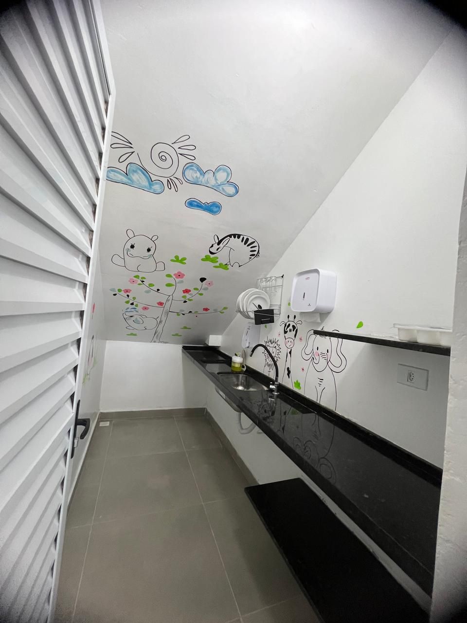 Uma cozinha estreita com paredes brancas, bancada preta e piso ladrilhado. Há desenhos de animais e flores na parede.