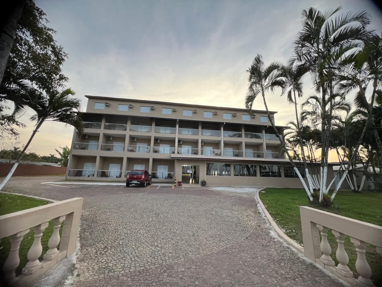 A multi-story hotel with a central entrance, a parking area, and surrounding palm trees.