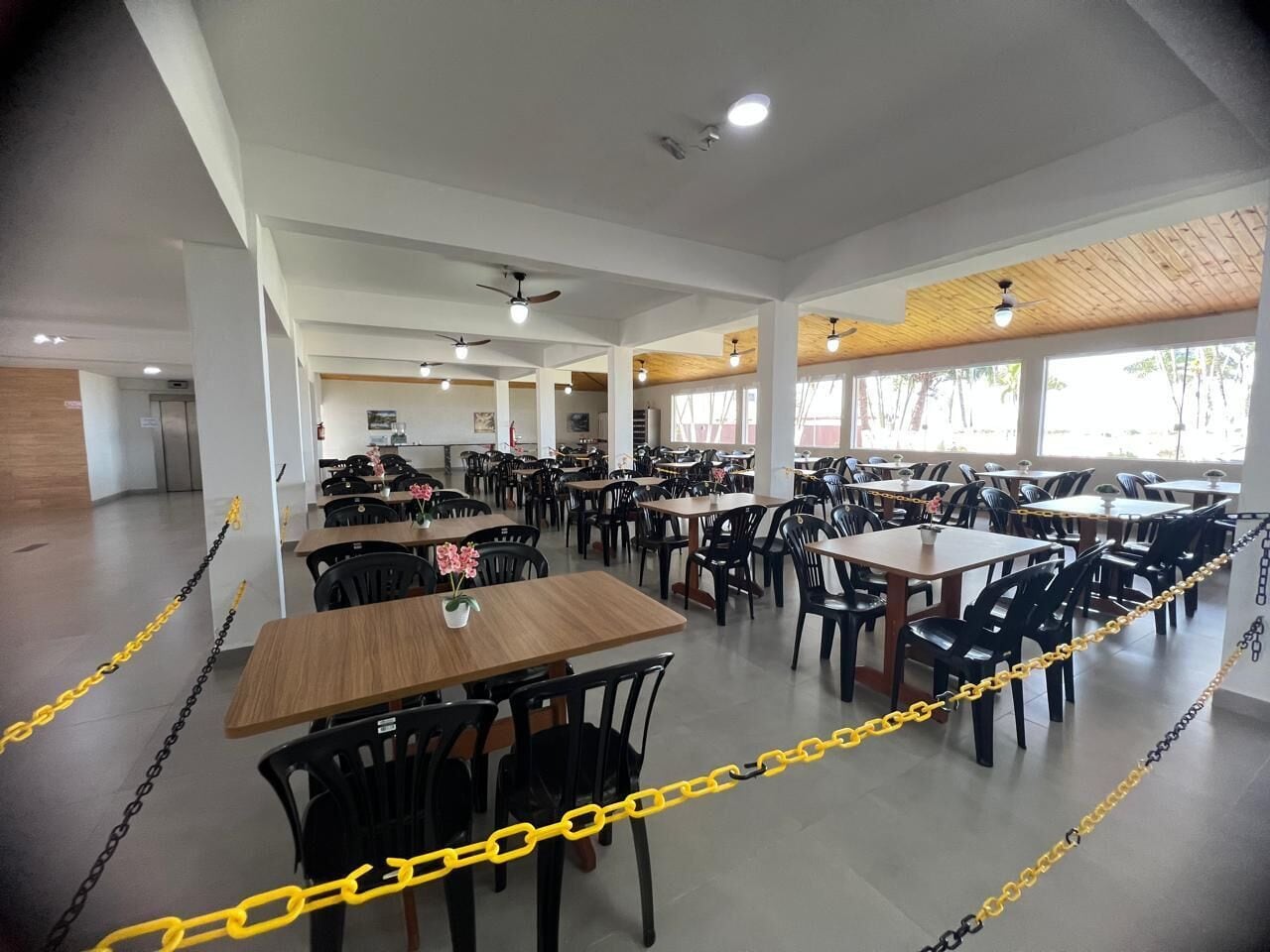 Un comedor amplio con varias mesas y sillas, delimitado por cadenas amarillas, y grandes ventanales que dejan entrar luz natural.