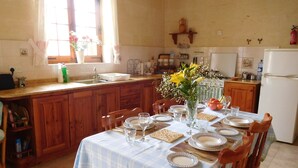 A kitchen with wooden cabinets, a dining table set with plates and glasses, and a window with flowers. - Dun Nastas Farmhouse (Sannat)