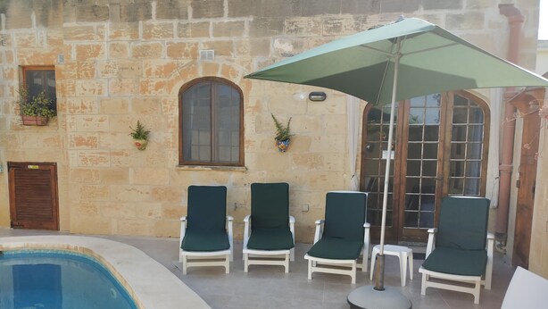 A poolside area with a stone wall, a wooden door, and a window. There are four lounge chairs and a table with a green umbrella. - Dun Nastas Farmhouse (Sannat)
