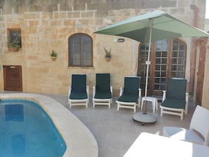 A poolside area with a stone wall, a wooden door, and a window. There are four lounge chairs and a table with a green umbrella.