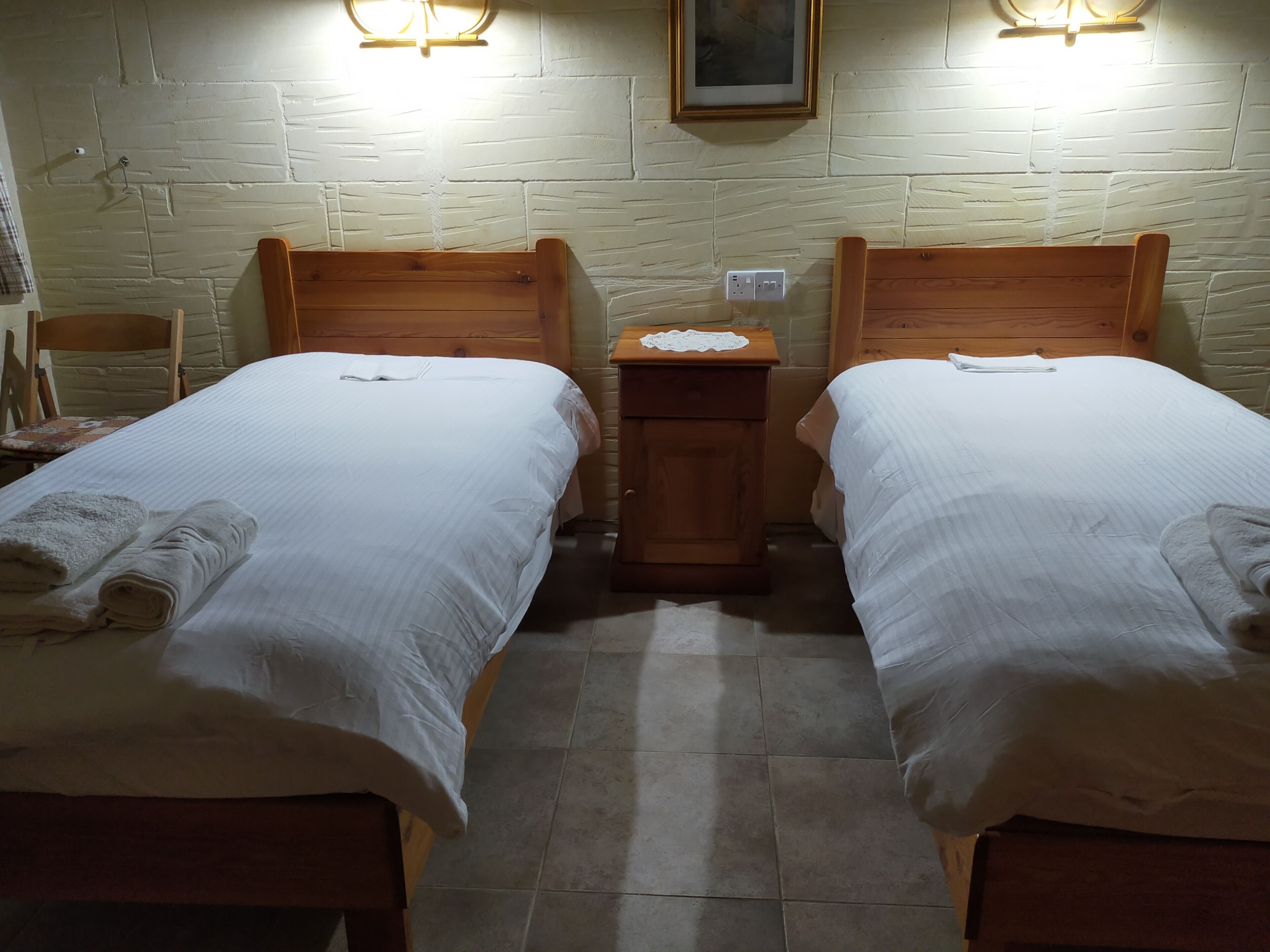 A room with two wooden beds, each with a white bedspread and rolled towels. There is a wooden nightstand between the beds with a small table and a framed picture above it. The walls are textured, and there is a light fixture on the wall.
