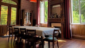 Ein Esszimmer mit großem Tisch, Stühlen, Kamin und großen, mit Vorhängen drapierten Fenstern.
