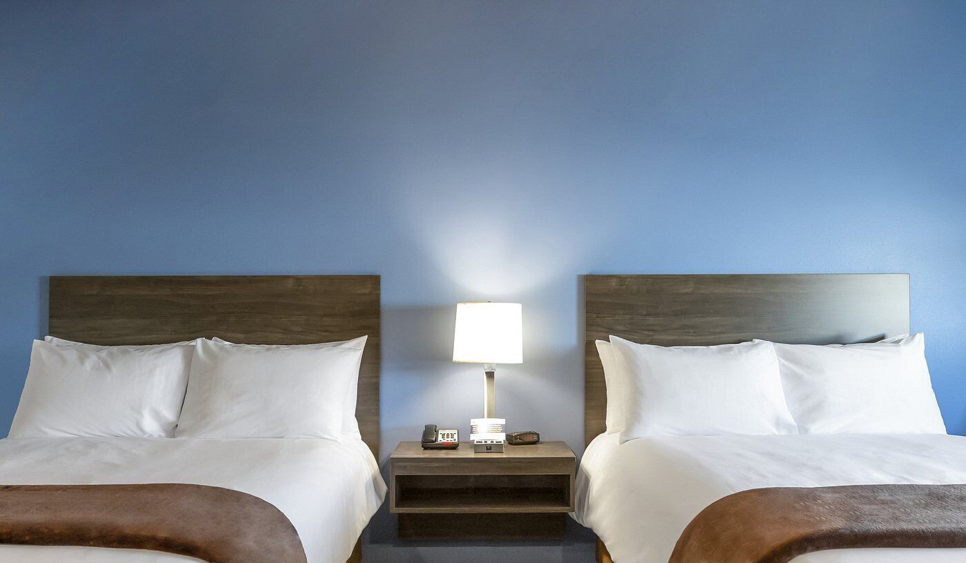 Two beds with white linens, a nightstand with a lamp, and a wooden headboard.