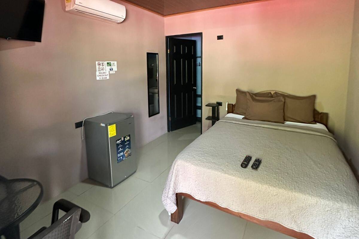 A hotel room with a bed, a desk, a chair, a TV, an air conditioner, and a trash bin.