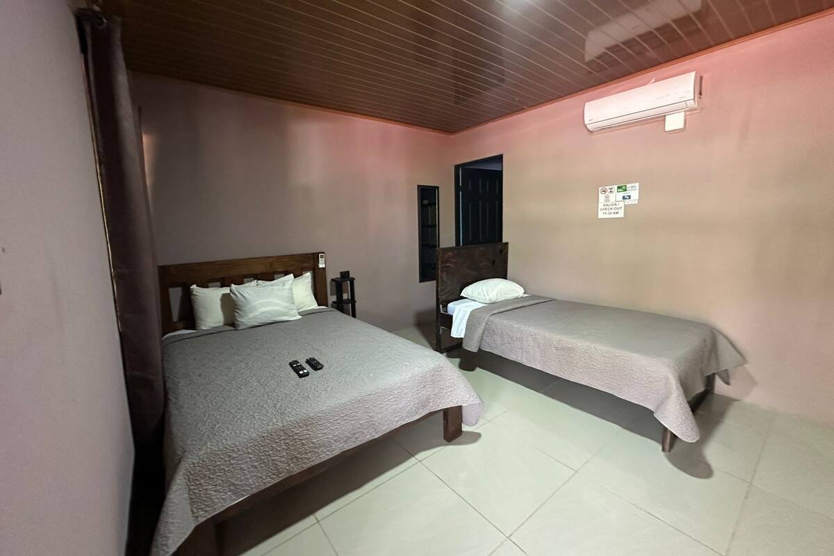 A room with two single beds, a wooden ceiling, an air conditioning unit, and a sign with check-in/out times.