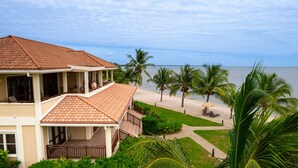 Una casa de dos pisos con techo de tejas, un balcón y vista a la playa con palmeras.
