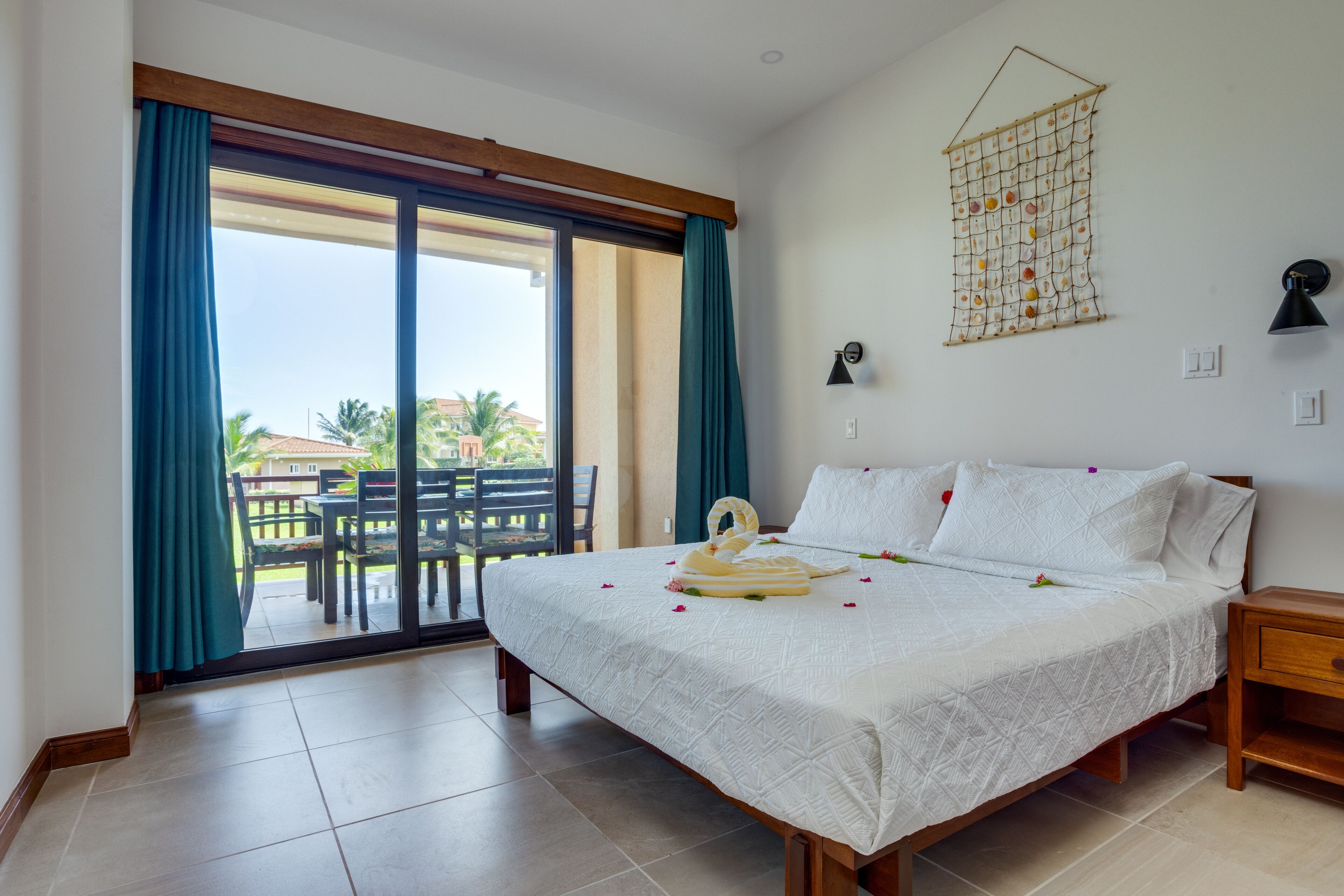 A hotel room with a large bed, a view of the outdoors, a wooden nightstand, and a wall-mounted lamp.