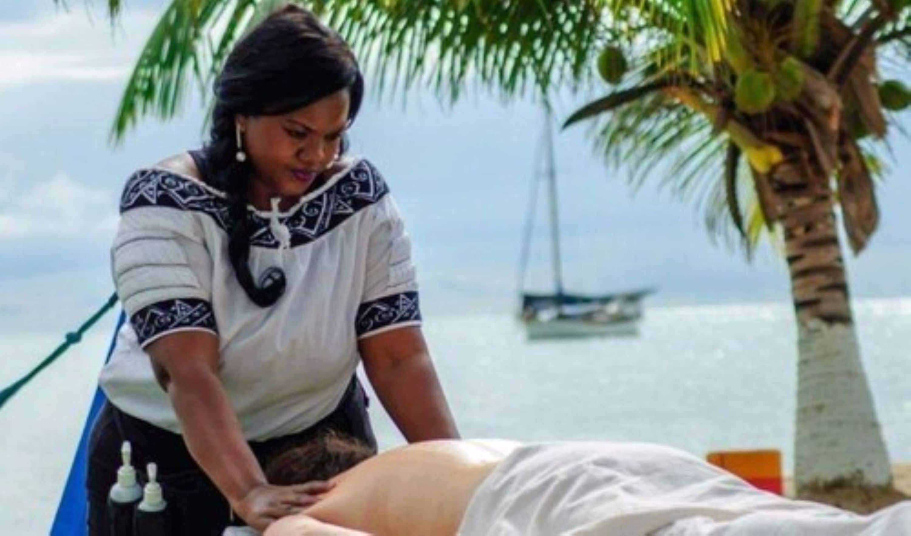 Uma pessoa recebendo massagem em uma praia, com uma palmeira e um barco ao fundo.
