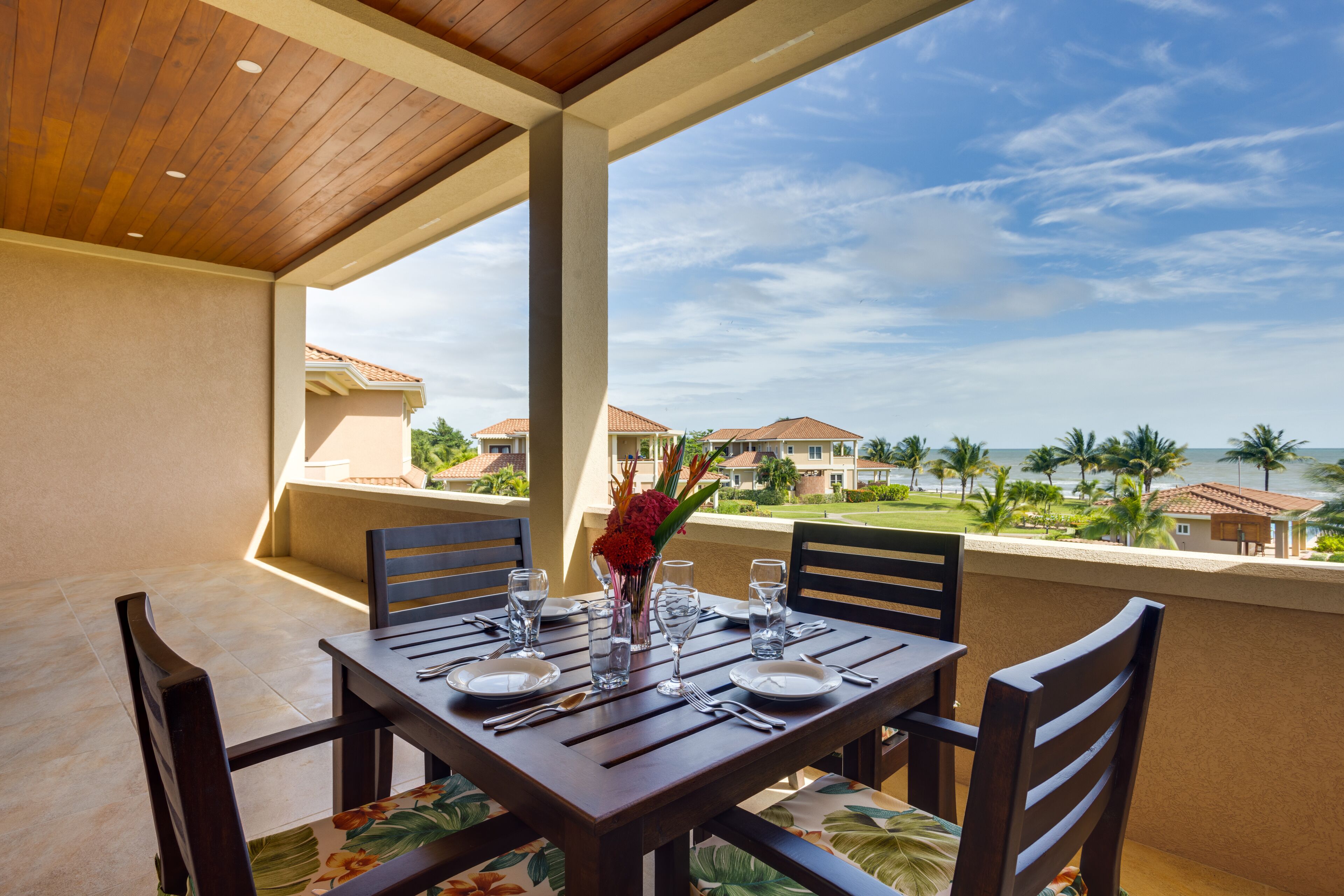 A dining table set for four with chairs, a vase with flowers, and a view of a coastal area with buildings and palm trees.