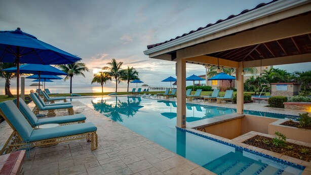 Zona de piscina con sillones de descanso y sombrillas, una vista despejada del atardecer y una superficie de agua tranquila que refleja el cielo.