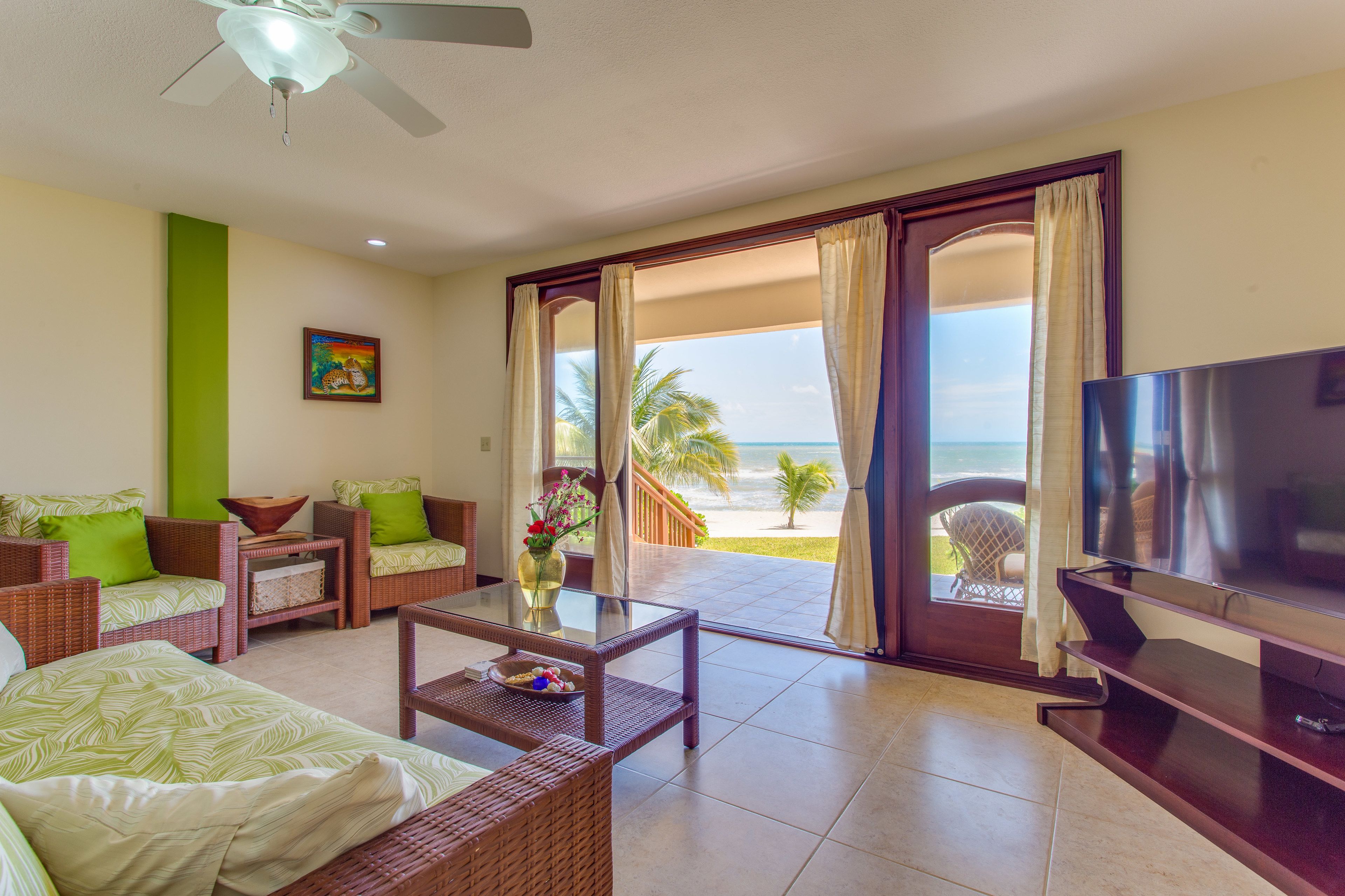 A room with a television, wicker furniture, a glass coffee table, and a view of the beach.