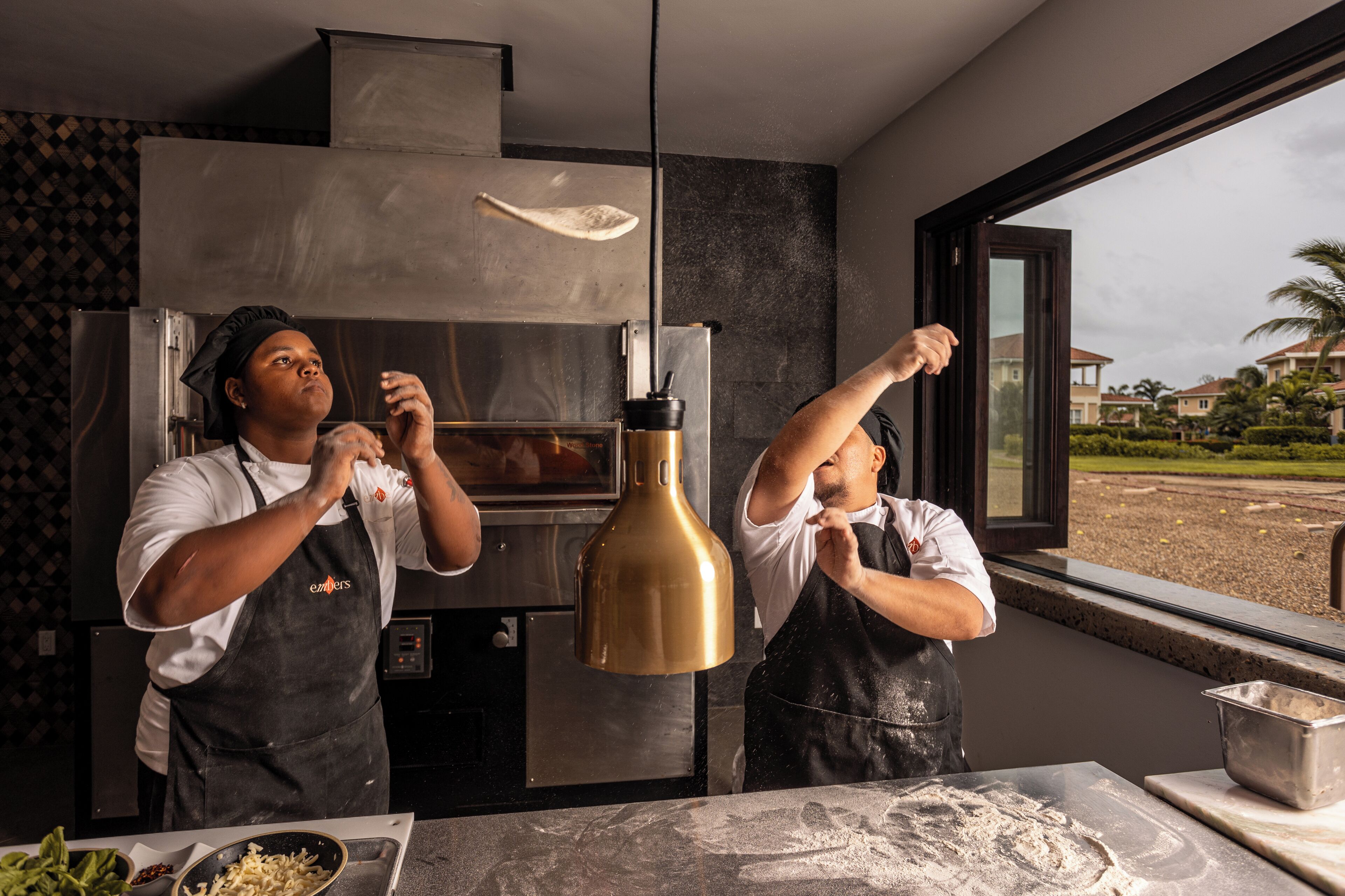 Two chefs in a kitchen with a modern design, a large oven, and a view of a residential area through the window.