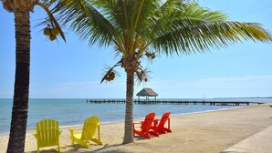 Una playa con sillas, una palmera y un muelle al fondo.