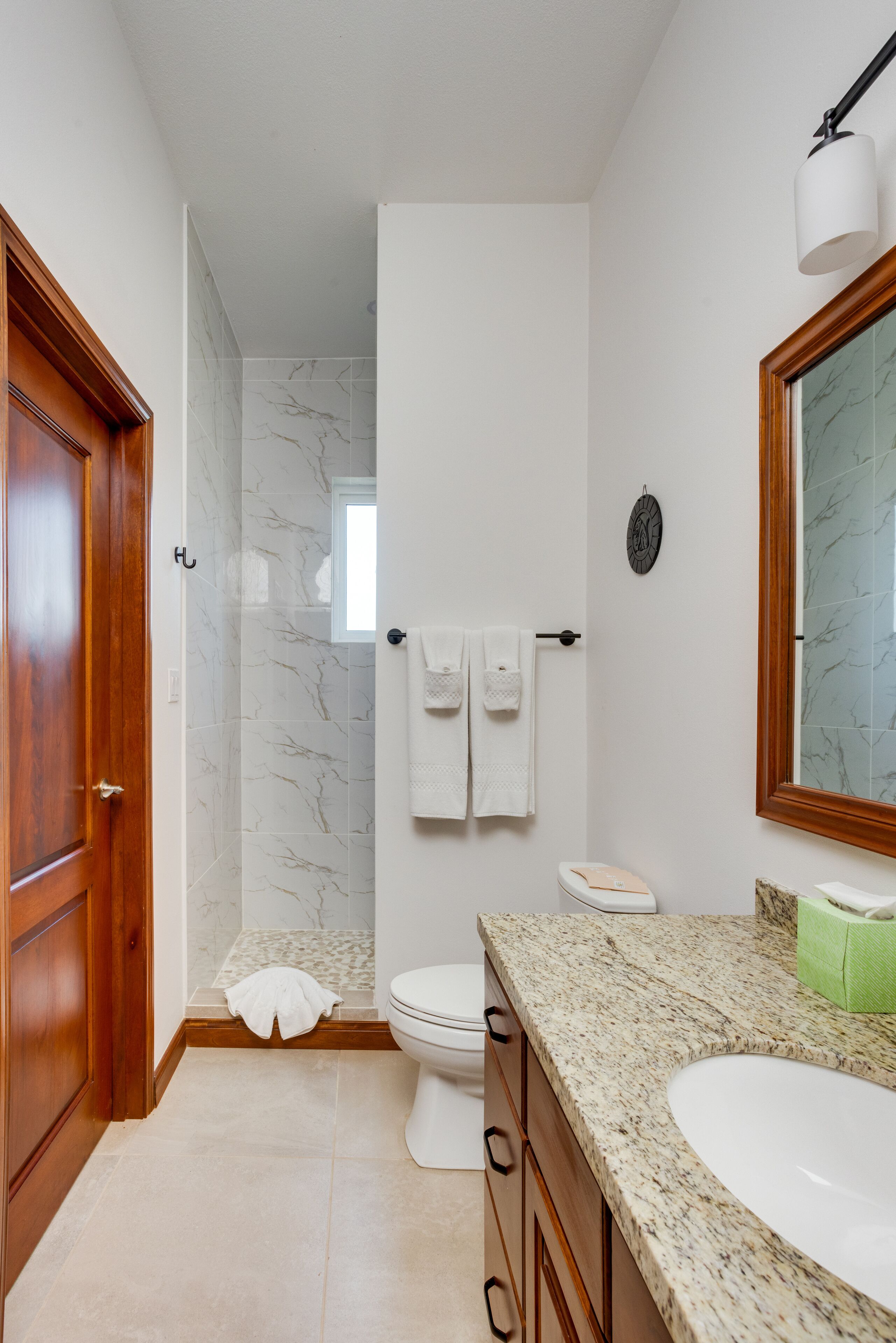 A bathroom with a wooden door, a shower with marble tiles, a toilet, and a sink with a granite countertop.