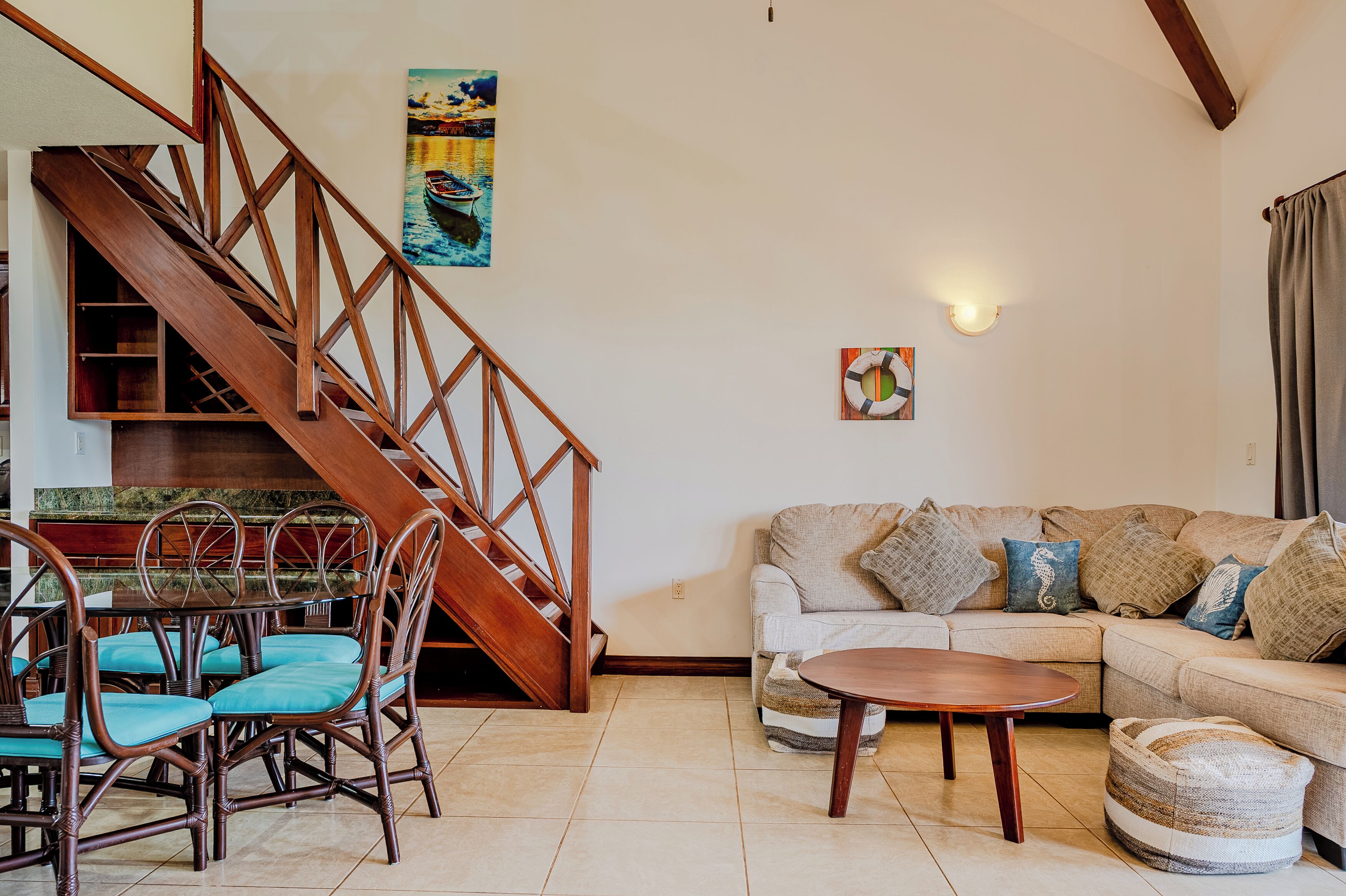 A living room with a wooden staircase, a dining table with chairs, a sofa with cushions, and a wall-mounted light fixture.