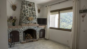 A stone fireplace with a television above it, a window with curtains, and a view of a forested area. - La Mariquilla, rural housing in Jubrique, Valle del Genal, natural paradise ... (Jubrique)