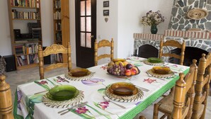 A dining room with a table set for four, surrounded by wooden chairs. A bookshelf filled with books is to the left, and a stone fireplace is to the right. - La Mariquilla, rural housing in Jubrique, Valle del Genal, natural paradise ... (Jubrique)