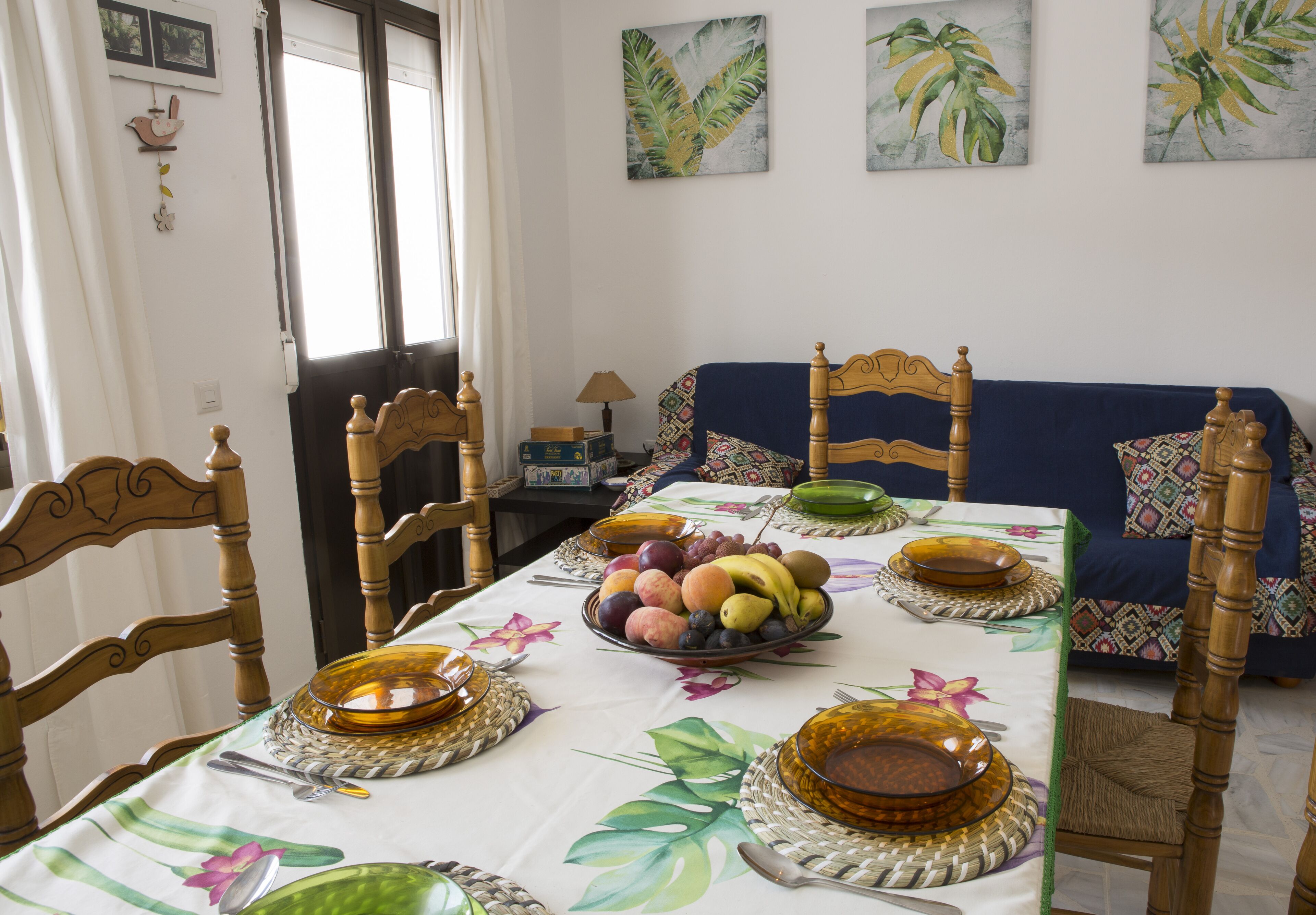 A dining table set with fruit and bowls, wooden chairs, a sofa, and artwork on the wall.