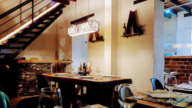 A restaurant with a rustic interior, featuring exposed brick walls, wooden beams, and a staircase. Tables are set with wine glasses and plates, indicating a dining area. - Coralite Hostel (Santa Marta)