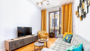 A modern living room with a flat-screen TV, a grey sofa with colorful cushions, a small round table, and a yellow armchair.
