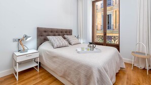 A bedroom with a bed, bedside table, lamp, and a window with a view of buildings.