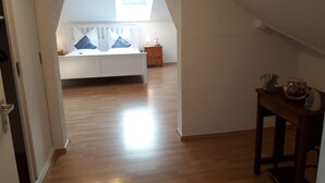 A spacious room with a high ceiling, a sloped roof, and a large window allowing natural light. A bed with pillows is positioned against the wall, and a small table with decorative items is on the right. - Large cozy apartment "Ziegenpeter" (DG = 2nd floor) - Kolvenbacher Ziegenhof 95sqm (Hohn)