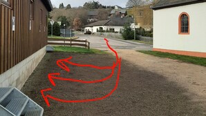 A gravel courtyard with a metal bench, a wooden building, and a white building with a red window. - Large cozy apartment "Ziegenpeter" (DG = 2nd floor) - Kolvenbacher Ziegenhof 95sqm (Hohn)